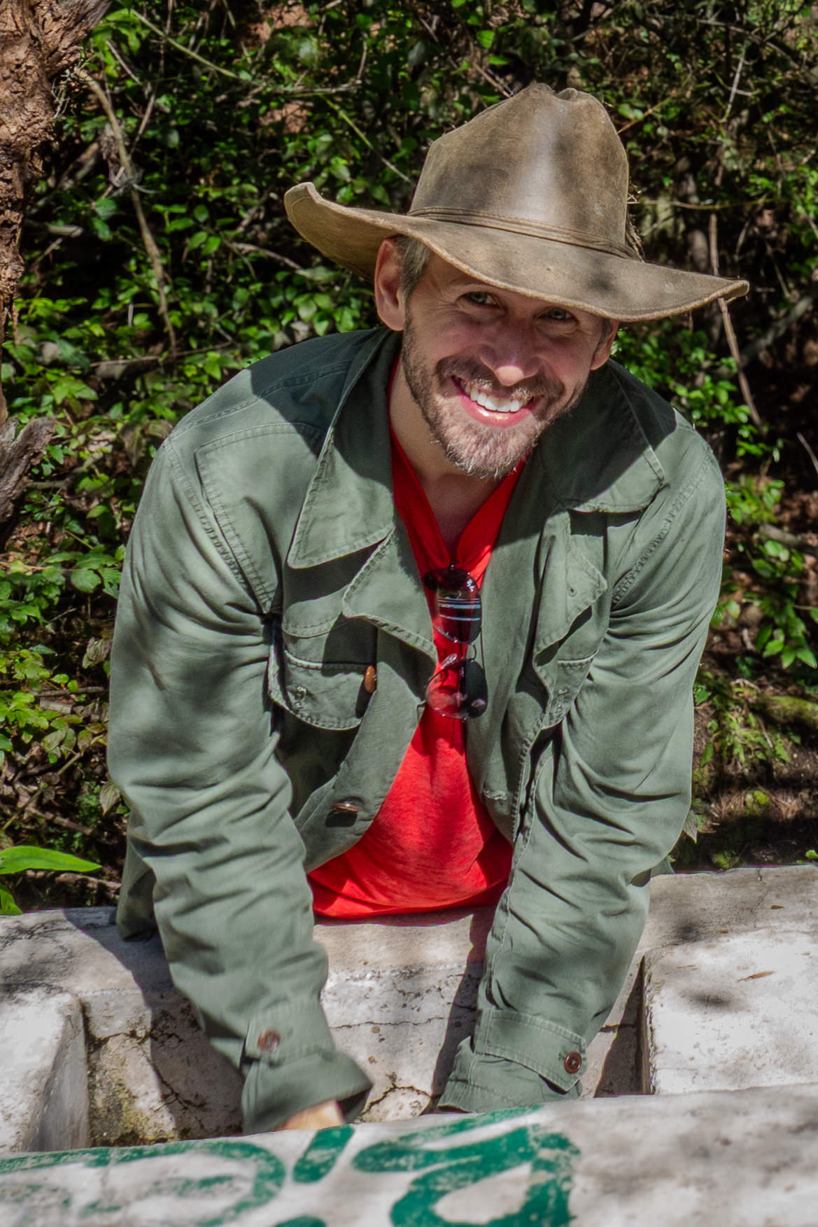 Man wearing a green jacket, red shirt, and brown hat standing outdoors with greenery in the background.