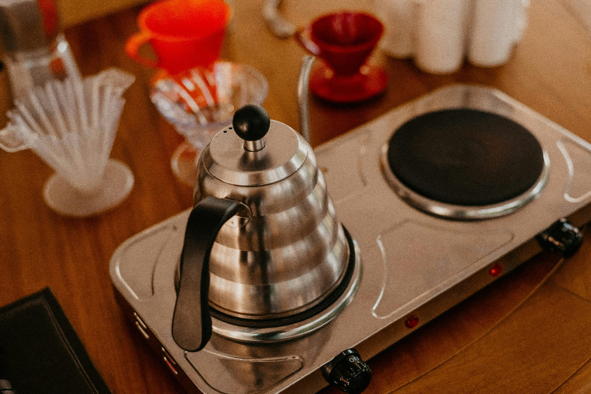Gooseneck kettle heating on a dual electric stovetop surrounded by pour-over coffee equipment on a wooden table.