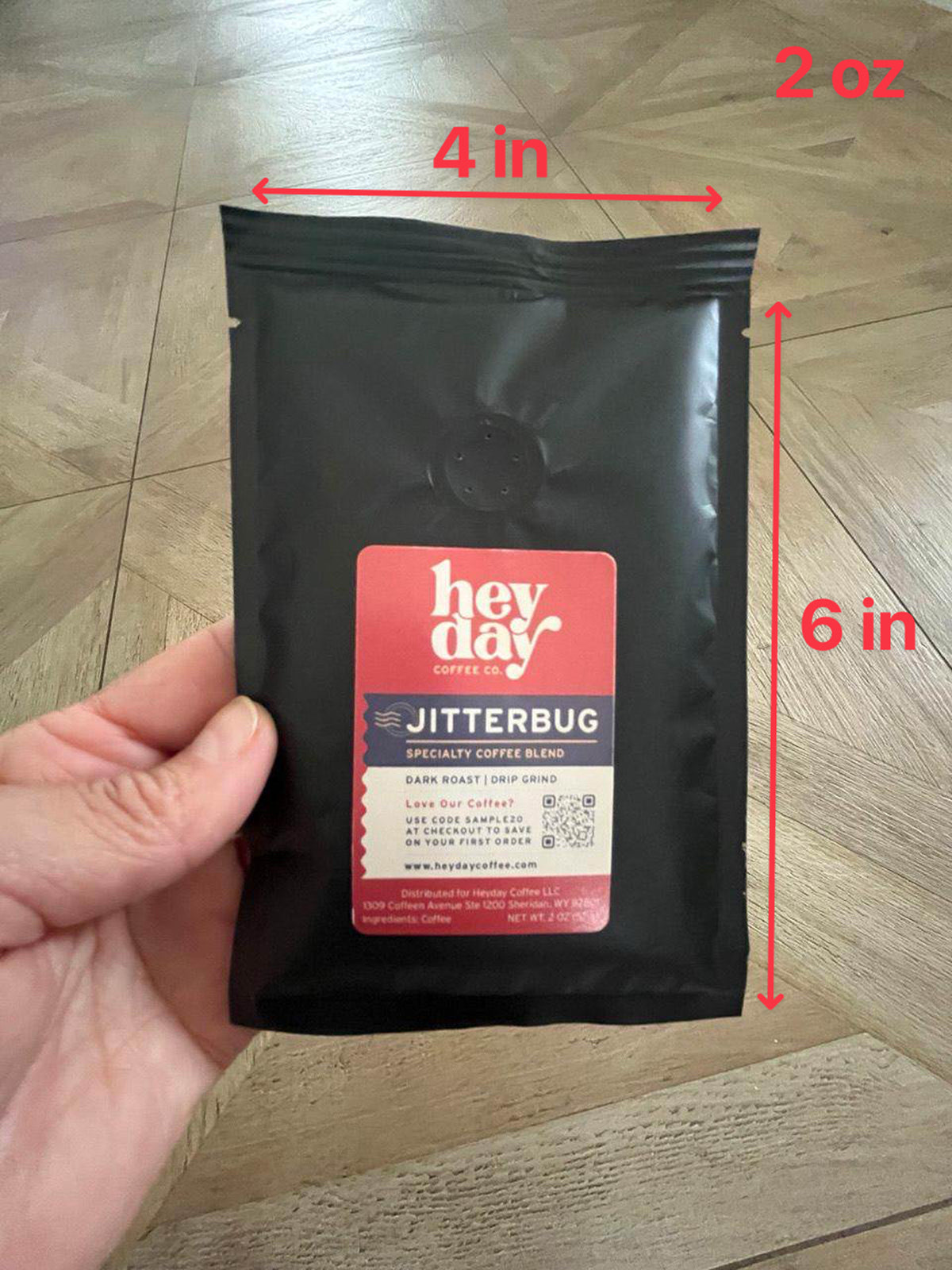 Hand holding a black coffee pouch above a wooden floor with annotated measurements